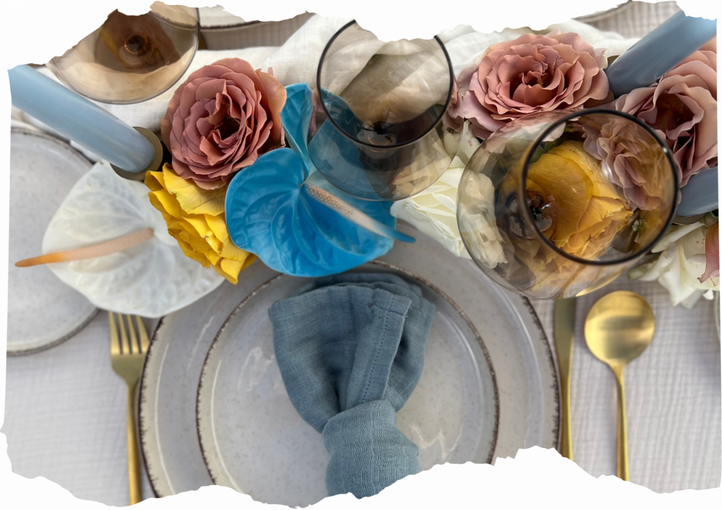 Vista detalle de plato sobre mesa decorada en tonos neutros y azules con rosas y anturios y una vela en forma de mano haciendo gesto de cuernos por Galana